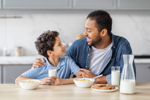 Happy african american father and preteen kid son have breakfast together, drinking milk, eating cereals and cooking. Dad hugging his child, have conversation while eating in kitchen at home - Photo, Image