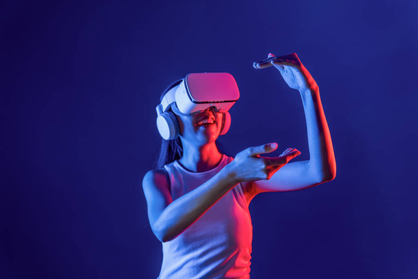 Inteligentna kobieta stojąca otoczona światłem neonowym nosi zestaw słuchawkowy VR łączący metawertyczną, futurystyczną technologię społeczności cyberprzestrzeni, wykorzystując obie ręce do interakcji z wygenerowanym obiektem wirtualnym. omamy. - Zdjęcie, obraz