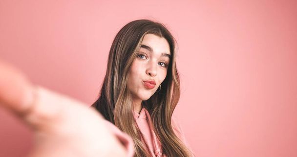 Pretty young female model with long blond hair puckering lips while looking at camera over pink background - Photo, image