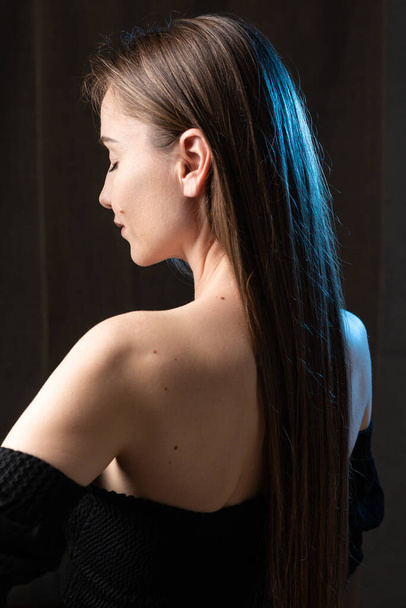Klasyczny portret studyjny młodej brunetki ubranej w czarny top, siedzącej na krześle na czarnym tle. - Zdjęcie, obraz