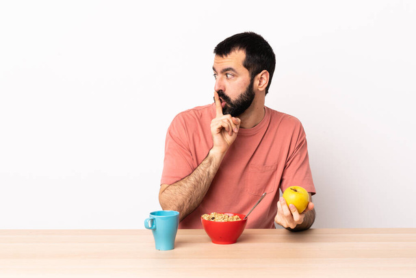 Кавказский мужчина завтракает в столе, показывая знак молчания жест положить палец в рот. - Фото, изображение