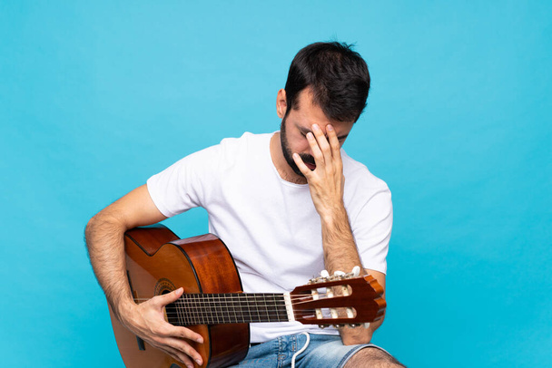 Jeune homme avec guitare sur fond bleu isolé avec expression fatiguée et malade - Photo, image