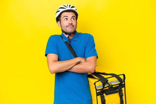 Junger gutaussehender Mann mit Thermorucksack über isoliertem gelbem Hintergrund macht eine Geste des Zweifels, während er die Schultern hebt - Foto, Bild