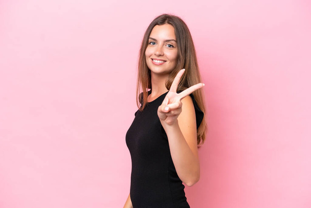 Junge kaukasische Frau auf rosa Hintergrund isoliert lächelnd und zeigt Siegeszeichen - Foto, Bild