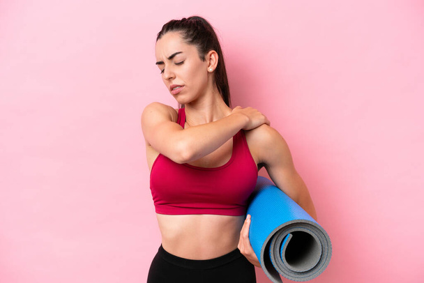 Junge Sportkaukasierin besucht Yoga-Kurse, während sie eine Matte auf rosa Hintergrund hält, die unter Schulterschmerzen leidet, weil sie sich angestrengt hat - Foto, Bild