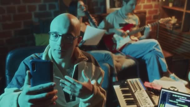 Panning footage of Caucasian bald blogger filming vlog on phone while his friends singing song to guitar on sofa in background in vintage cozy studio - Footage, Video
