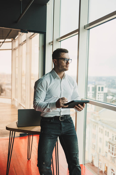 Ένας όμορφος άντρας με γυαλιά και ένα πουκάμισο στέκεται στο γραφείο στο τραπέζι και δουλεύει σε ένα τάμπλετ. Ένας σύγχρονος χώρος εργασίας με χώρο γραφείων - Φωτογραφία, εικόνα