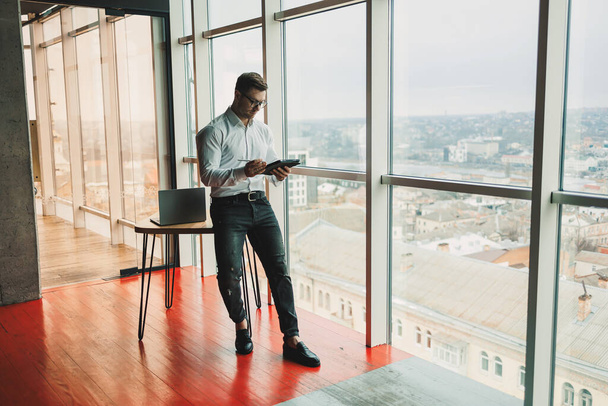 Egy jóképű férfi szemüvegben és ingben áll az asztalnál az irodában, és egy táblagépen dolgozik. Modern munkaterület irodahelyiséggel - Fotó, kép