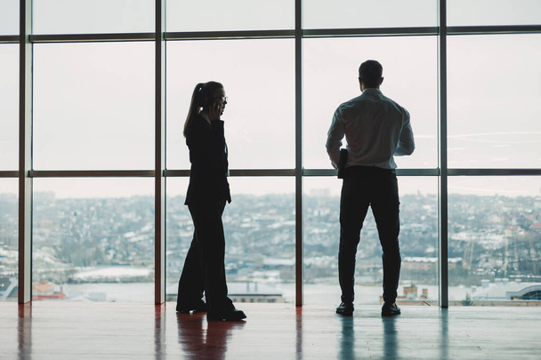 Мужчина и женщина стоят в кабинете на фоне большого панорамного окна. Современное просторное рабочее пространство с сотрудниками. Бизнес партнеры - Фото, изображение