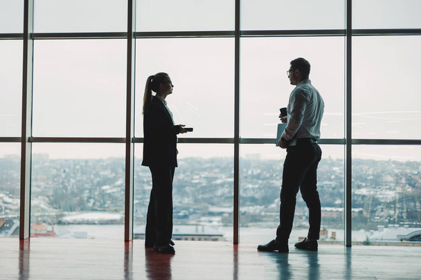 Vor dem Hintergrund eines großen Panoramafensters stehen ein Mann und eine Frau im Büro. Moderner, großzügiger Arbeitsplatz mit Mitarbeitern. Geschäftspartner - Foto, Bild