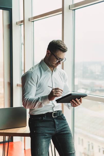U stolu stojí pohledný muž v brýlích a košili a pracuje na tabletu. Moderní pracovní prostor s kancelářskými prostory - Fotografie, Obrázek