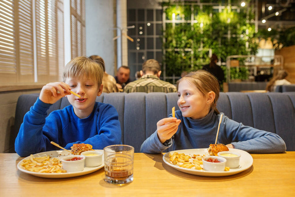 Мальчик и девочка в голубых топах делятся игривым моментом во время еды гамбургеров и картошки фри в ресторане. - Фото, изображение