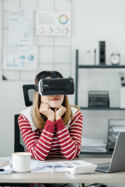 Kontemplativní mladá žena se sluchátky VR sedí na svém pracovišti a zkoumá virtuální prostředí. - Fotografie, Obrázek