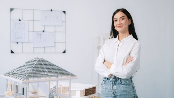 Close up portrait of young beautiful professional architect engineer cross arms and poses confidently with house model at white modern office with blueprint. Creative living and design. Immaculate. - Photo, Image