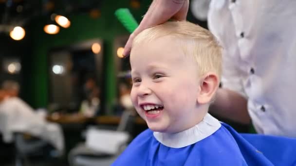 A férfi fodrász fésülködéssel és ollóval vágja a kisfiú haját. Közelkép boldog gyermek kap hajvágás felnőtt férfi, valószínűleg borbély. Professzionális fodrász és aranyos ügyfél a modern fodrászatban. - Felvétel, videó
