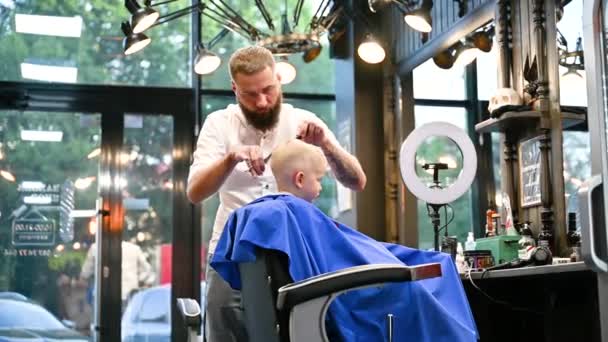Peluquero hombre cortar el pelo de los niños pequeños usando peine y tijeras. A un niño le cortan el pelo hombres adultos, probablemente peluquero. Peluquería profesional y cliente lindo en la barbería moderna. - Metraje, vídeo
