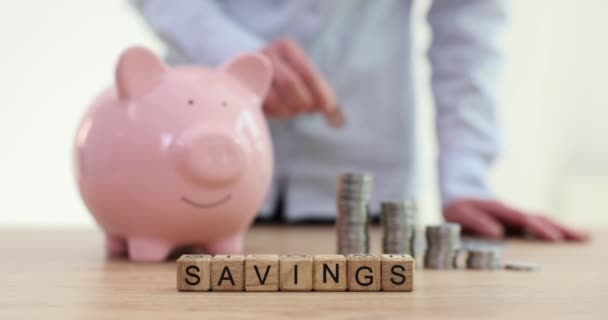 Word Savings on wooden cubes and woman throwing coins into piggy bank on table. Lady keeps money for future expenses. Family budget - Footage, Video
