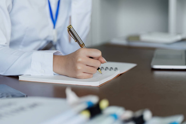 Egy nő ír egy jegyzetfüzetbe egy tollal. Fehér inget visel és kék a nyaka. A jelenet professzionális környezetet sugall. - Fotó, kép