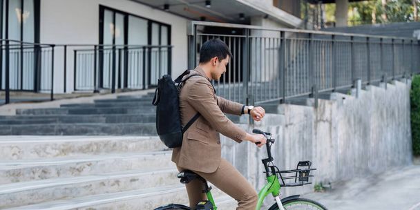 Mężczyzna w garniturze jeździ na zielonym rowerze z koszykiem. Nosi zegarek i plecak. - Zdjęcie, obraz