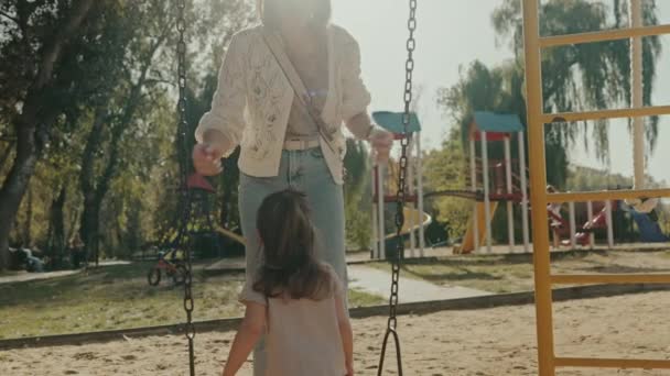 Mother holding her daughter and placing her on the swing in the park. Nature, weekend and mom push little girl in playground for relaxing, childhood and - Footage, Video
