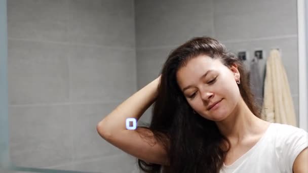 Junge Frau mit dunklem lockigem Haar bei einer Selbsthaarmassage mit Kopfhautmassager oder Haarbürste zur Stimulierung des Haarwuchses im heimischen Badezimmer. Reflektierter Blick auf den Spiegel. Hochwertiges Foto - Filmmaterial, Video