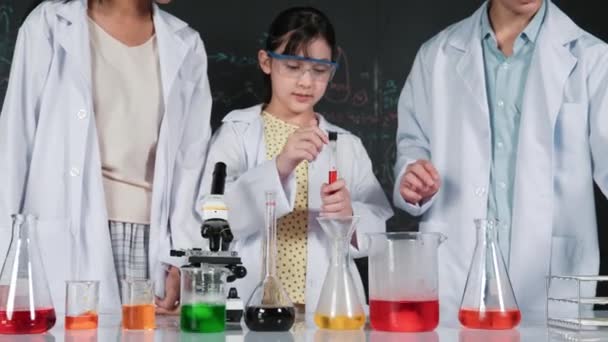 Chica de la escuela mezcla solución de color mientras que el apoyo académico de los estudiantes haciendo experimento en la mesa con microscopio y líquido. Estudiante diverso de pie en pizarra con la teoría de la ciencia escrita. Edificación - Metraje, vídeo