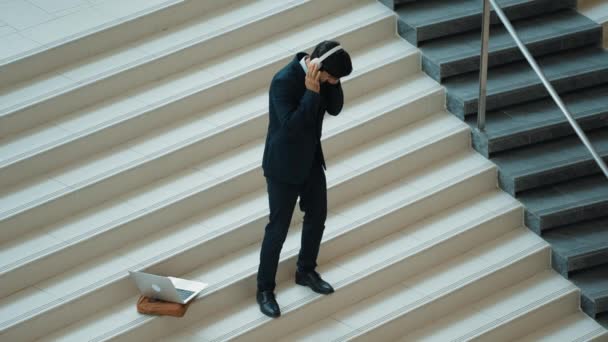 Professzionális üzletember áll a lépcsőházban, miközben a fejhallgatóról zenél. Képzett projektmenedzser táncol, miközben hallgat pihenni hang laptoppal elhelyezett lépcsőn. Modern tánc. Eredményes. - Felvétel, videó