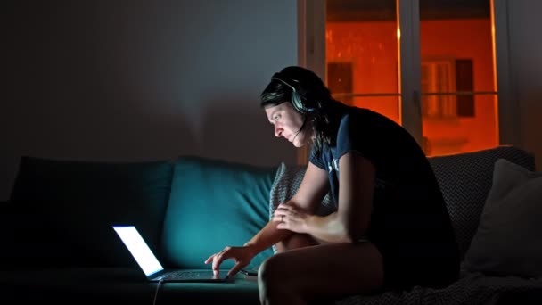 女性は夜にラップトップを使用しています リビングルームで - ライトオフとヘッドフォンでソファーに座って - 映像、動画