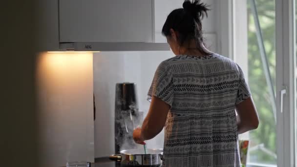 Vrouw bereidt maaltijd op inductie kachel - roestvrij stalen pot emitting stoom als ze beweegt, natuurlijk licht uit het appartement venster. Casual binnenlandse dagelijkse Candid Scene - Video