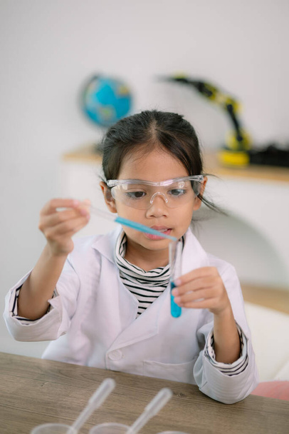 Μικρό χαριτωμένο κορίτσι με μικροσκόπιο που κρατάει ένα εργαστηριακό μπουκάλι με πειράματα νερού μελετά επιστήμονες στο σχολείο. Εκπαιδευτική επιστημονική έννοια. - Φωτογραφία, εικόνα