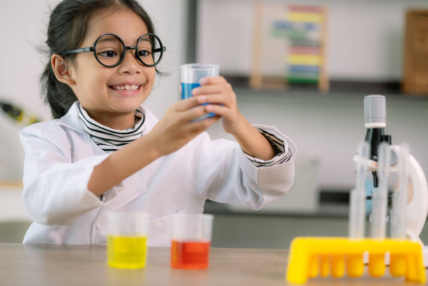 Μικρό χαριτωμένο κορίτσι με μικροσκόπιο που κρατάει ένα εργαστηριακό μπουκάλι με πειράματα νερού μελετά επιστήμονες στο σχολείο. Εκπαιδευτική επιστημονική έννοια. - Φωτογραφία, εικόνα
