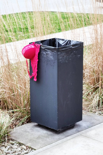 A black trash can is shown with a red hat placed on top of it. The contrast between the black and red colors is striking, drawing attention to the unusual combination of objects. - Photo, Image