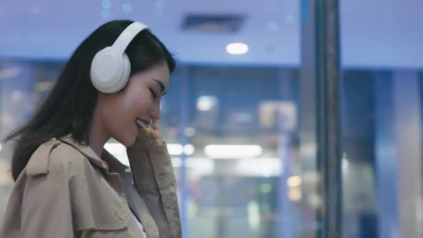 アジアのイブニングモールを歩いている間,ヘッドフォンで踊る楽しい魅力的な少女. チャーミングな若い女性は,オンラインで音楽を聴くために電話でアプリケーションを使用しています. 人と音楽のコンセプト - 映像、動画