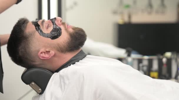Fryzjer nakłada czarną maskę nawilżającą na twarz mężczyzny podczas golenia. Dermatolog nakłada czarną maskę na twarz brodatego pacjenta, aby usunąć pory i zmarszczki wideo 4k. - Materiał filmowy, wideo