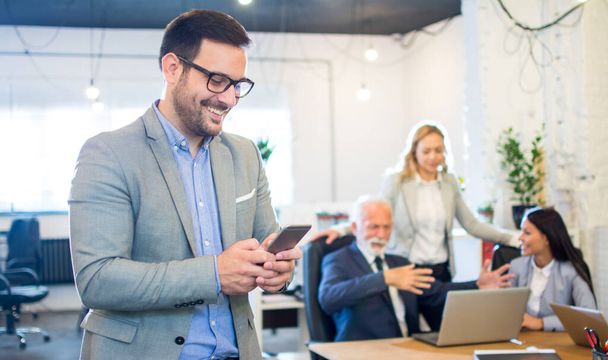 Smiling businessman using phone in the office with colleagues in the background - Photo, Image