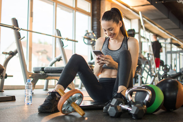 Piękna dziewczyna fitness siedzi i relaksuje się na macie treningowej i przy użyciu telefonu w siłowni. - Zdjęcie, obraz