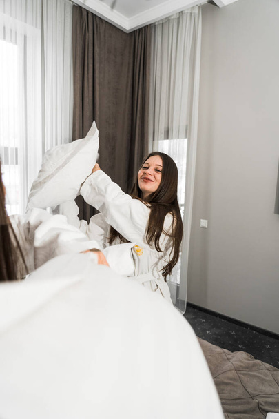 Two young women in bathrobes laughing and hitting pillows in bed. Hotel holiday concept - Photo, Image