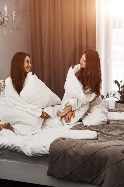 Dwie młode kobiety w szlafrokach śmiejące się i uderzające w poduszki w łóżku. Koncepcja wakacji - Zdjęcie, obraz