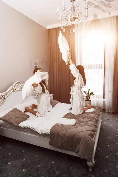 Bornozlu iki genç kadın yatakta gülüyor ve yastıklara vuruyor. Otel tatil konsepti - Fotoğraf, Görsel