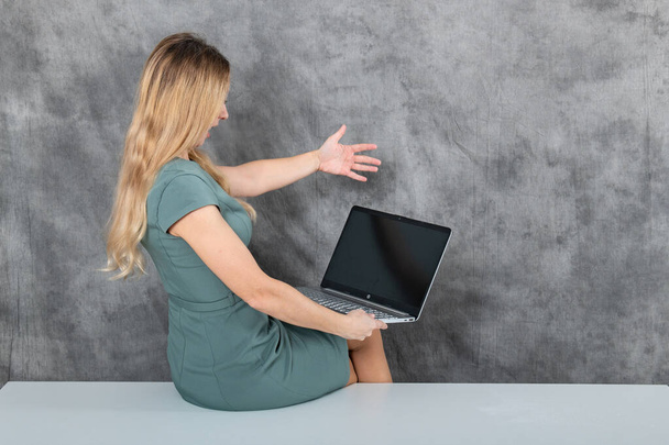 Eine junge blonde Frau sitzt zu Hause auf einem Holztisch und arbeitet an einem Laptop. Sie ist lässig gekleidet und konzentriert sich auf ihre Arbeit und präsentiert das Konzept der Fernarbeit und Flexibilität. - Foto, Bild