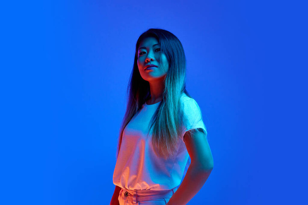Selbstbewusste, schöne Asiatin, Geschäftsfrau, die vor blauem Hintergrund im Neonlicht vor der Kamera posiert. Geschäftskonzept, Unternehmertum, menschliche Emotionen, Selbstdarstellung. - Foto, Bild