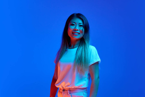 Sorrindo positivo jovem, bela mulher asiática vestida roupa casual posando olhando para a câmera contra fundo gradiente azul em luz de néon. Conceito de emoções humanas, auto-expressão, diversão e alegria. - Foto, Imagem