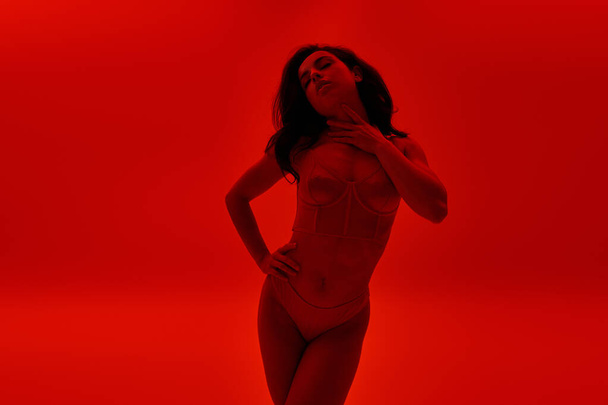 Junge Frau steht selbstbewusst in Dessous vor leuchtend rotem Hintergrund. - Foto, Bild