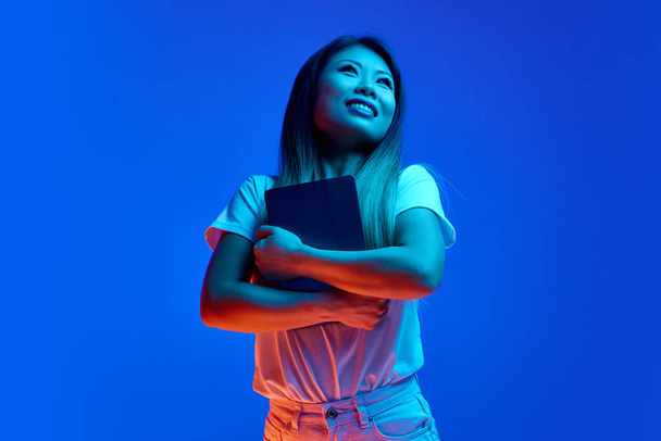 Jovem, bela e alegre mulher asiática segurando tablet digital e olhando para o fundo do gradiente azul em luz de néon. Conceito de negócio, estudo e trabalho à distância, inicialização, vendas, compras. - Foto, Imagem