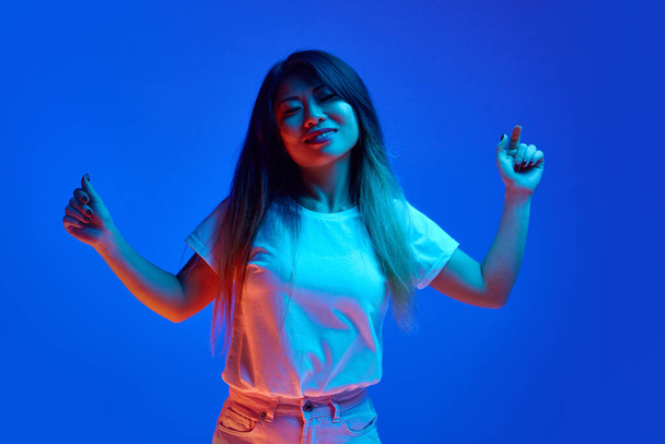 Overgelukkig, ontspannen Aziatische vrouw dansend hand in hand tegen blauwe gradiënt studio achtergrond in neon licht. Concept van menselijke emoties, zelfexpressie, muziek en dans, plezier en vreugde. - Foto, afbeelding