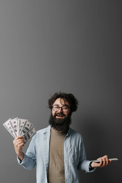 Un hombre vestido casualmente se levanta contra un telón de fondo gris neutro. Él sostiene una pila de dinero en efectivo en una mano y un teléfono inteligente en la otra, lo que sugiere una transacción o concepto de finanzas digitales. - Foto, Imagen