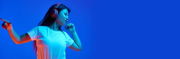 バナー。 ネオンライトの青い勾配の背景に対してヘッドホンで音楽を聞くアジアの女性. テキストを挿入する負のスペース. 人間の感情,自己表現,ダンス,音楽の概念,楽しさ,喜び. - 写真・画像