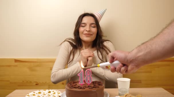 The girl is sitting in front of a table with a festive cake, in which a candle in the form of the number 18 is burning, which she blows out. Birthday celebration concept. - Footage, Video