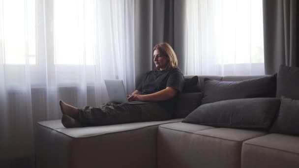 Happy young caucasian woman using texting mobile phone or chat and smiling with laptop computer sitting of the sofa in home. Communication female looking message at cell phone or smartphone. Slow - Кадры, видео