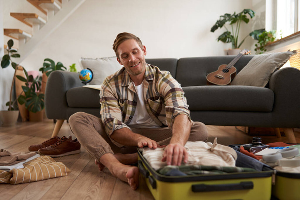 Image of young man, traveller, sitting on floor with opened suitcase, packing clothes for a trip, going on holiday. Tourism concept - Photo, Image